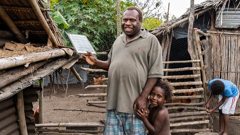 Man smiling, with a child by his side, holding a small solar panel in his hands 