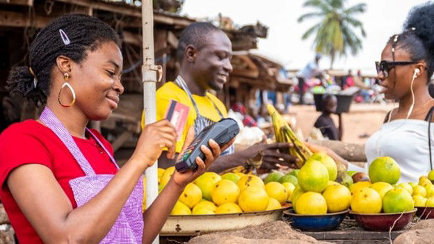 An African woman uses a card reader to facilitate digital transactions for her fruits business at the local marketplace
