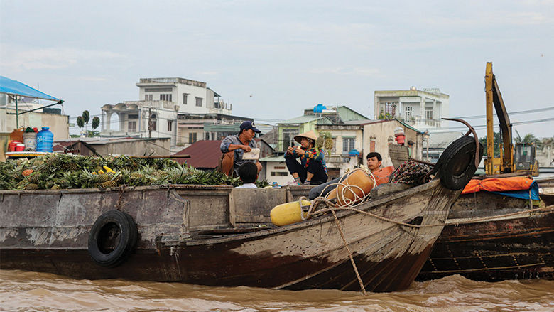 A wooden boat loaded with produce travels along a muddy river, with several people sitting and talking on board. Houses and s