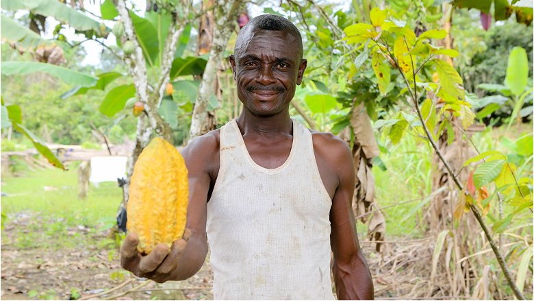 Male farmer from Ghana smiling at the camera, outdoors, holding a yellow cocoa pod in his hand