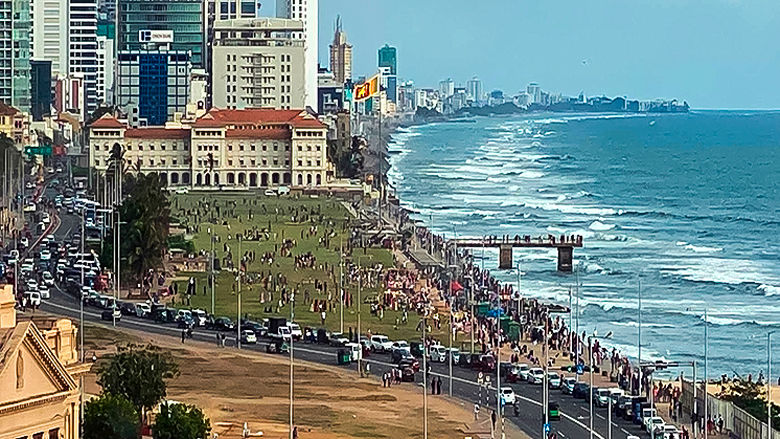 A zoomed out image of a coastal community in Sri Lanka. Multiple buildings are seen, as well as people in a seaside park.