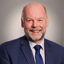 Male with beard smiling, wearing navy blue suit and tie and light grey blouse