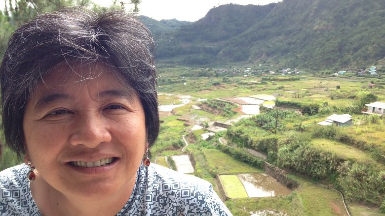 Joan Carling smiling, wearing a blue and white patterned dress. In the background, a lush valley with houses and bushes.
