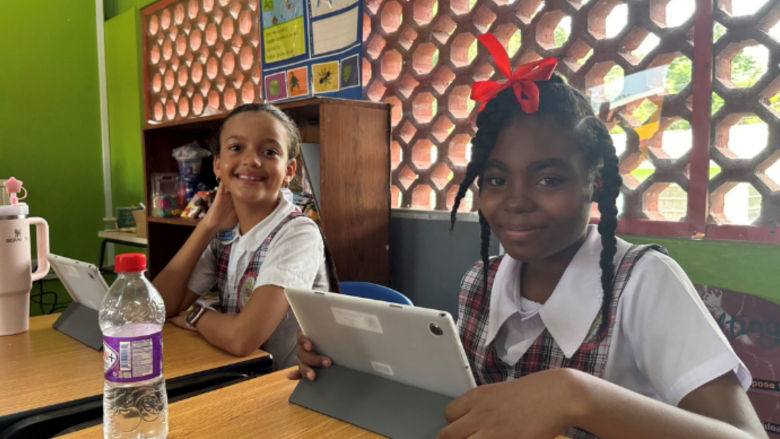 Two young girls from St Lucia sitting at their desks in class smile while holding their tablets