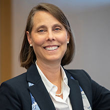 Female with short brown hair smiling, wearing a white blouse, dark blue suit and a pearl necklace and earrings 