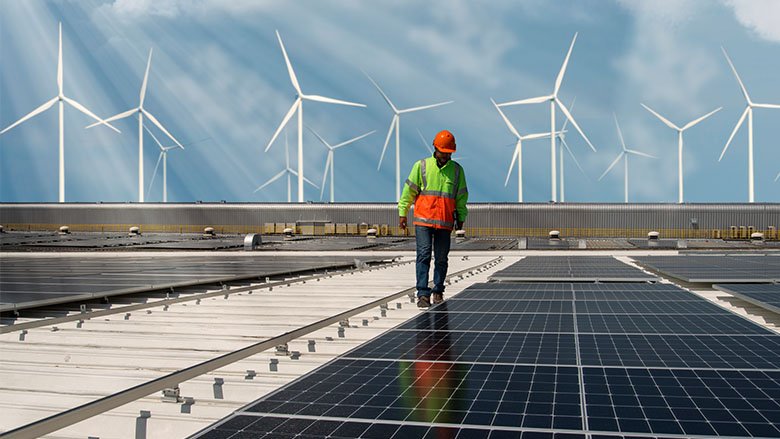 An engineer walks next to some solar panels. In the background there are some windmills