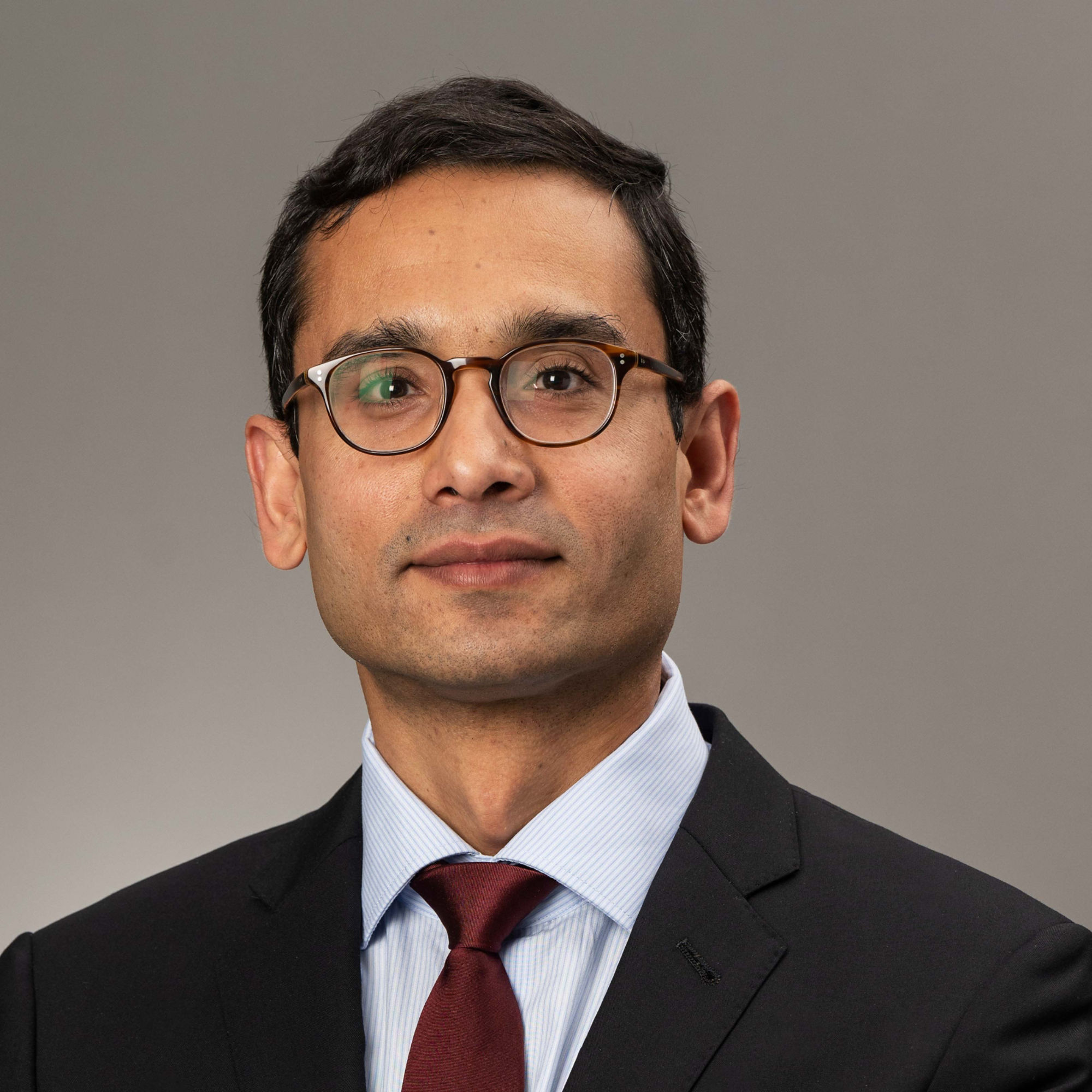 Male with short brown hair wearing eyeglasses, black blazer, striped white and blue blouse and dark red tie