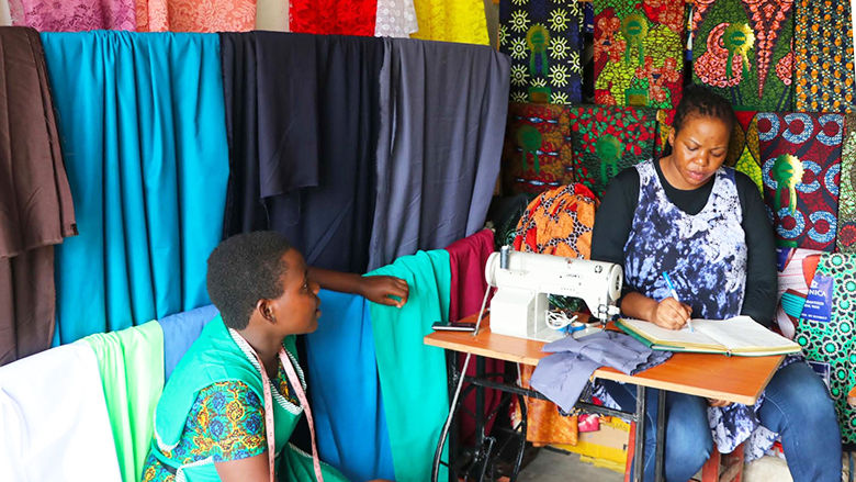 One lady sitting in front of a sewing machine with another watching