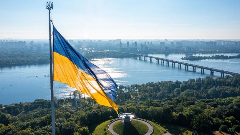 Ukraine flag on a sunny day, river and bridge behind in the background.