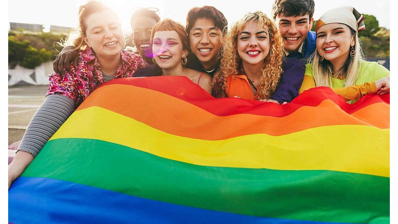 Group of friends, smiling, prop up a LGBTI flag, with the colors red, orange, yellow, green, and blue visible.