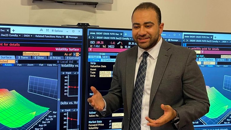 Hatem presenting in front of monitors with financial data