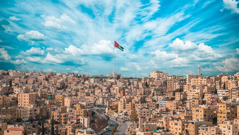 The Jordan flag  appears at full mast at the center of the image, on a cloudy day, amidst a landscape of building's in Amman.