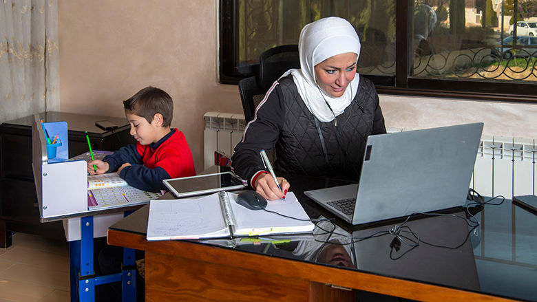 Jordanian woman works at computer with nearby child doing schoolwork 