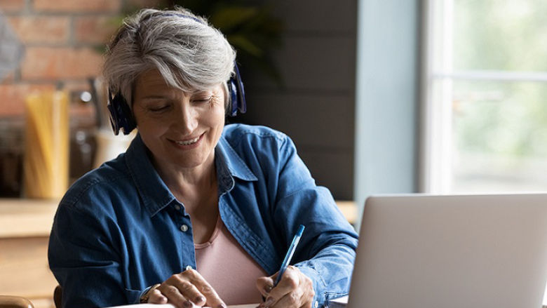 women with grey hair writing in notebook sitting in front of the laptop
