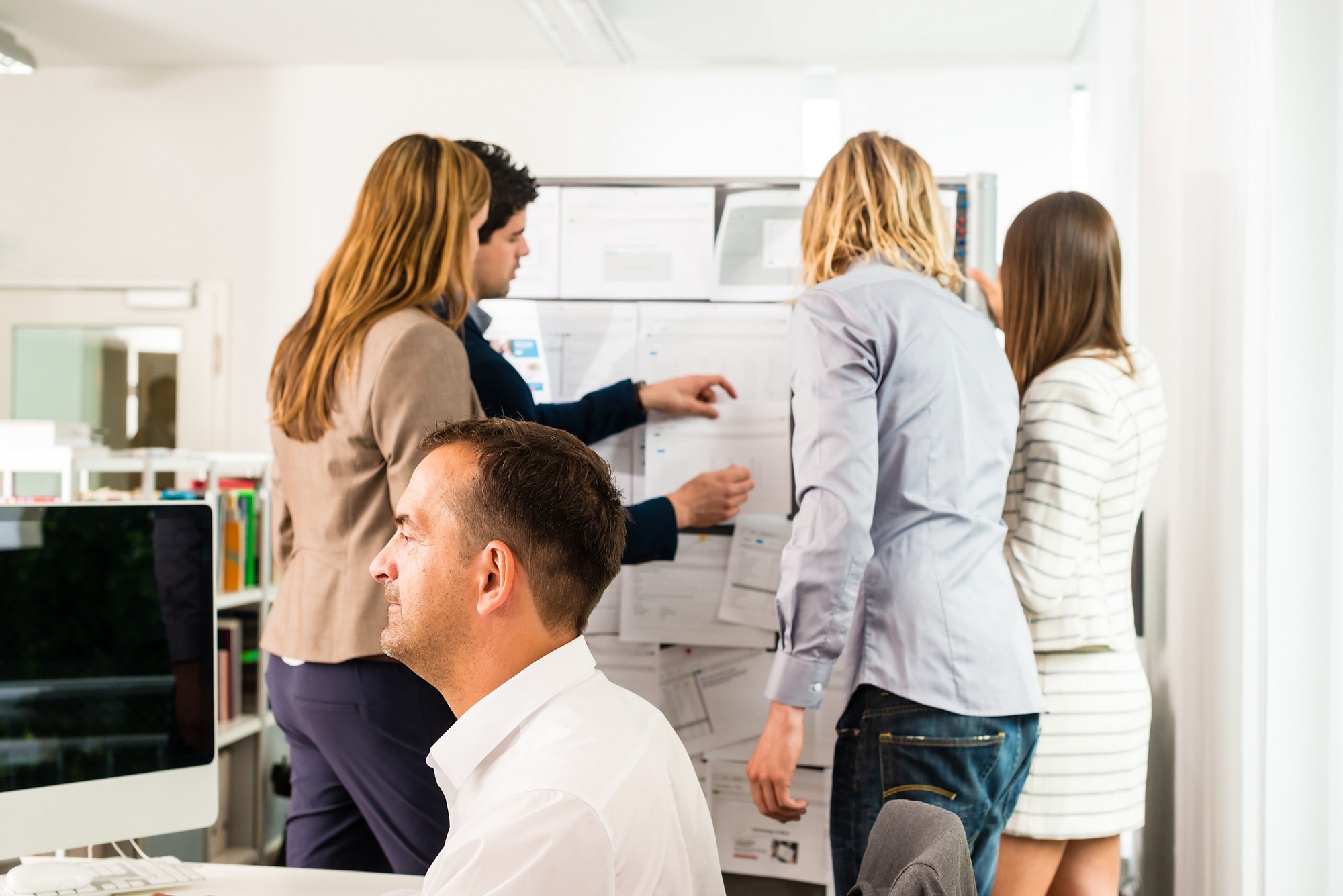 Businesspeople working in the office - some of them looking at bulletin board and discussing designs pinned at it, another guy in the front is working with several computer screens. Mixed caucasian