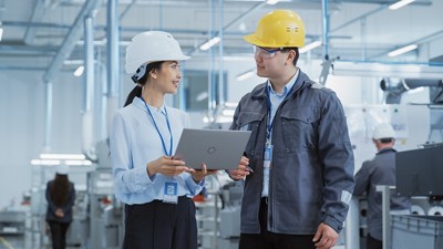 Two Diverse Young Heavy Industry Engineers in Hard Hats Walking with Laptop Computer and Talking in a Factory. Two Manufacturing Employees at Work in Research and Development Facility.