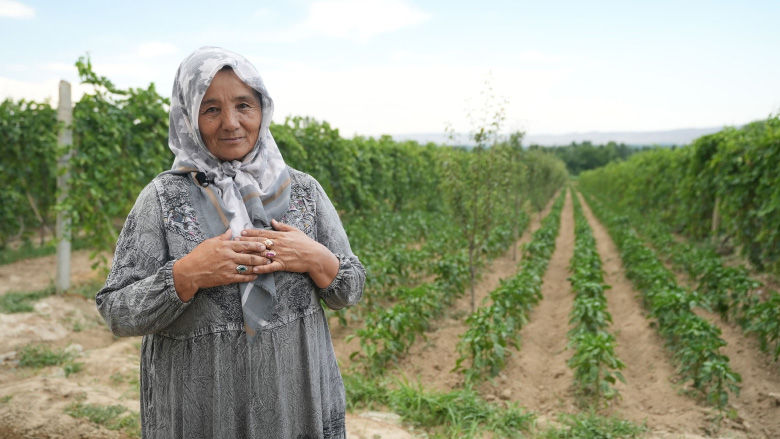 A woman stands by rows of potatoes and grapevines